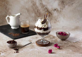 Chocolate trifle with whipped cream cheese in a glass, individual layered dessert decorated with piece of chocolate bar and biscuit sticks. Biscuit crumbs, coffee and dried small roses on the table