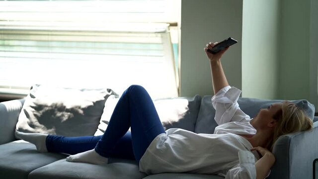 blonde woman is taking selfie by smartphone, lying on couch at home, day at weekend