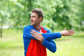 Front view of a sportsman stretching hands while standing outdoor in a park and looking by side.