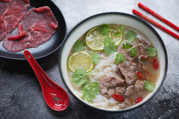 Pho bo soup served with slices of raw beef meat over black and white stone background, elevated view