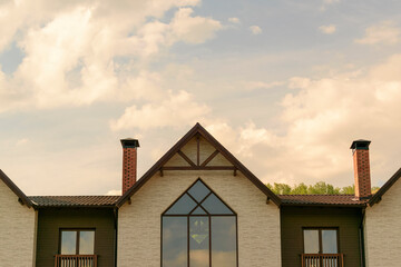 Attic townhouse. Roof with brown tiles. Fireplace pipes. Glazed windows. Background - trees, sky with clouds. Concept of modern low-rise housing construction, home purchase.