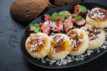 Cheesecakes or syrniki served with jam, coconut flakes and strawberries on a black plate, close-up, selective focus