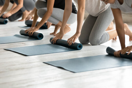 Close up young diverse barefoot people folding sport floor mats together, finishing yoga class lesson indoors, group of athletic friends unrolling rubber carpets, starting yoga workout at gym studio.
