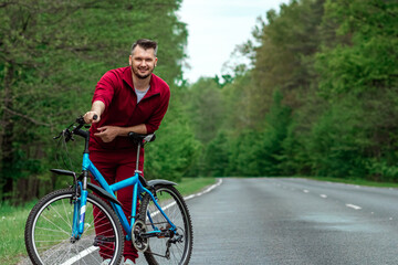 Naklejka premium A man in a tracksuit stands next to a bicycle on a road in the forest. The concept of a healthy lifestyle, cardio training. Copyspace.