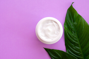 Soft white cream in jar with bright green leaves on pink background with copy space. Spa treatment and procedure at home.