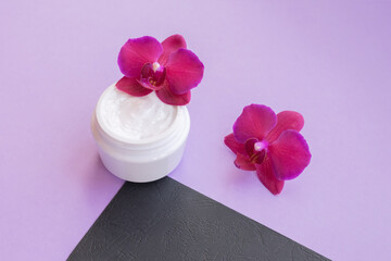 White container with cream for face and body with three magenta colored orchid flowers on purple and black background. Concept of delicate or eco friendly cosmetics