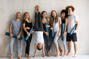 Fototapeta na wymiar Sportive fit smiling indian ethnicity woman standing upside down on hands near wall with young happy excited multiracial people in activewear holding floor mats, warming up before training indoors.