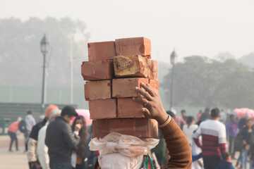 Image of female worker carrying bricks over her head on a construction site at crowdy public place
