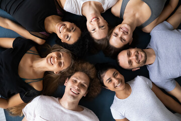 Head shot close up young attractive mixed race happy people in sportswear lying on floor in circle, relaxing after intensive workout or enjoying beak pause time together indoors, teambuilding concept.