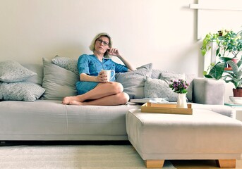 40+ woman sitting on a gray sofa in a denim dress and drinking tea