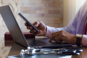 Male doctor work on laptop computer and using mobile smart phone , search application for medical knowledge with medical stethoscope  on the desk at clinic or hospital . Teleconference, EMR concept.