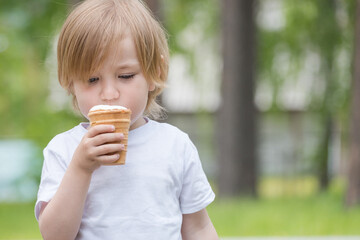 Beautiful baby with blue eyes in white tshirt is eating glass of white ice cream.