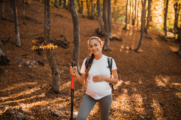 Portrait of young happy pregnant woman nordic walking in autumn forest with backpack and trekking sticks. Healthy, wellness and active lifestyle in maternity time.