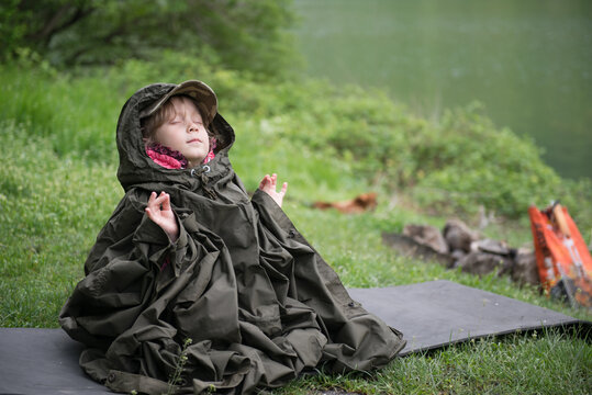 A child sits on the lake shore on a tourist Mat in the rain in a raincoat tent and meditates. An image with selective focus.
