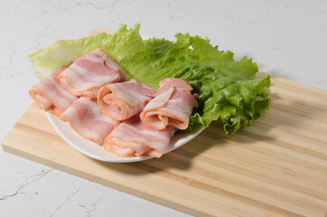 Rolled bacon slices are placed on a lettuce leaf on a plate. Bamboo cutting Board and texture table as background.
