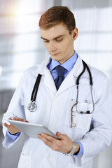 Young doctor man is standing in a sunny hospital office, holding a computer tablet in his hands. Perfect medical service in clinic. Happy future in medicine and healthcare