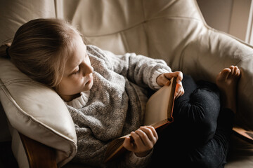 A blonde caucasian girl sitting curled up on a chair reading a book
