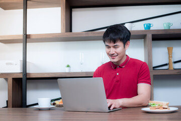 A young Asian man sat and worked from home in the dining room. Which is decorated in wood and looks modern, he is using a laptop while drinking coffee and eating sandwiches.