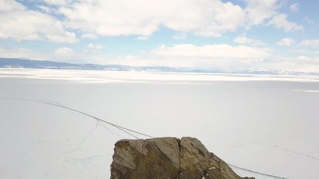 Steep rock and the shore of a frozen lake. Clip. Aerial view of the steep cliff and mountains covered by bald trees and the lake under the ice and snow, winter nature landscape.