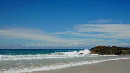 Seascape: surf breaking on rocks, with sandy beach in the foreground, clouds on the horizon, and blue sky above.