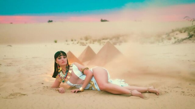 fantasy woman Egyptian pharaoh Cleopatra lies on sand in sunset desert. Pyramids, dunes, sandstorm, hurricane. Historical costume, queen clothes, white dress. Gold Crown necklace jewelry, accessories