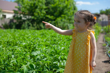 A little girl in a yellow dress is standing in the garden. Pointing to the side.