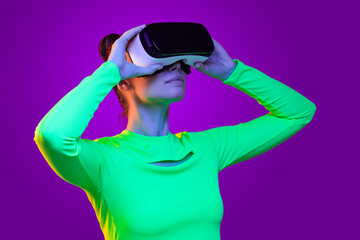 Neon cyberpunk virtual reality concept. Young  girl in green  top, wearing VR headset, holding it with both hands, isolated on purple background