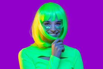 Happy teen girl dressed in green neon top, glitter on cheeks, smiling and laughing at party, isolated on purple background