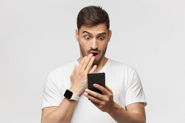 Young man in white t-shirt looking at phone screen with shocked face, isolated on gray background