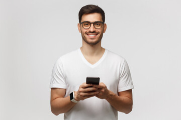 Portrait of young handsome man in white t-shirt and eyeglasses, standing with phone in hands,...