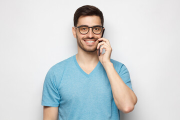 Portrait of young handsome man wearing blue tshirt and glasses, talking on the phone, holding smartphone next to ear, answering call, isolated on gray background