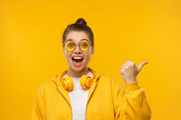 Young excited surprised girl in pointing right to copy space with thumb isolated on yellow...