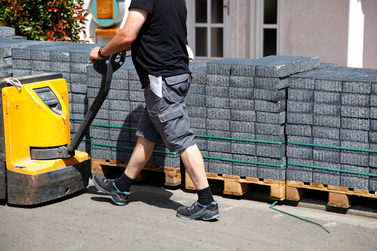 A fork pallet truck stacker with stack of concrete paving slabs. delivery man with pallet jack, delivering of concrete paver blocks on wooden pallets on the construction site. side view. delivery note