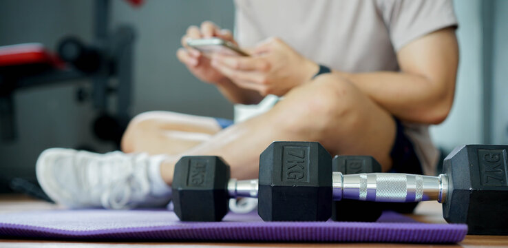close up on dumbbell on the floor with man sitting and playing smartphone to watching or learning from online course personal trainer program for workout at home concept