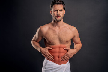 Fototapeta premium Handsome young bearded man isolated. Cropped image of shirtless muscular man is standing on gray background. Man holding his stomach. Experiencing stomach pain.