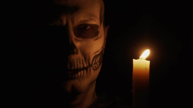 A man with skeletal makeup looks at the camera. Man casts spells. Skeleton in the dim light of a candle. Halloween and horror concept.