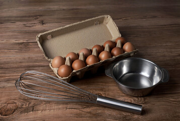 Raw chicken eggs in a cardboard box with stainless steel bowl and metal whisk on brown wooden background.