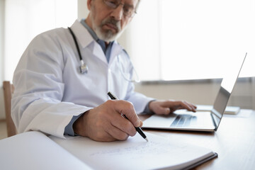 Close up of elderly grey-haired male doctor or GP sit at desk in hospital write in paper medical journal work on laptop, serious old mature physician make notes fill patient anamnesis in book register - obrazy, fototapety, plakaty