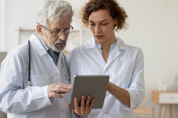 Concentrated man and woman doctors in white medical uniforms look at tablet screen discuss patient anamnesis or checkup results together, diverse GP talk brainstorm make decision using pad gadget - obrazy, fototapety, plakaty
