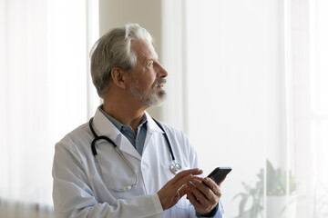 Pensive elderly male doctor in white medical uniform use cellphone look in distance window thinking pondering, thoughtful mature man GP or physician consult client online on smartphone, make decision