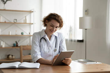 Smiling female nurse or GP in white medical uniform sit at desk in hospital or clinic work on...