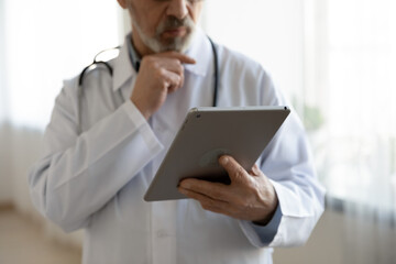 Crop close up of mature male doctor physician in medical uniform look at tablet screen make decision, pensive man GP or therapist work use modern pan gadget in hospital, medicine technology concept