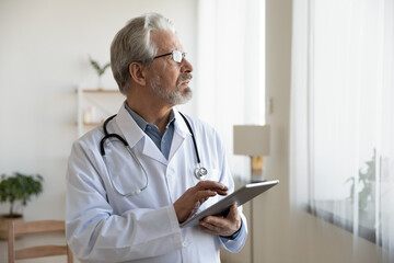 Pensive elderly male doctor in white medical uniform work on modern tablet look in distance...