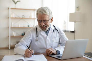 Smiling old male doctor in glasses and white uniform sit at desk in hospital work on laptop write in journal, happy mature senior man physician fill patient medical history anamnesis in paper register - obrazy, fototapety, plakaty