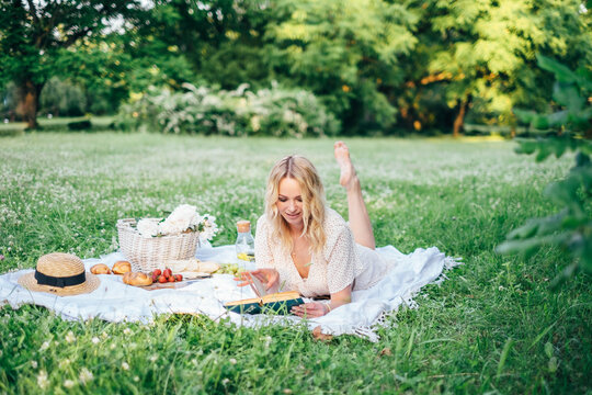 Young beautiful european blonde longhaired girl in white dress at nature.Spends the weekend outside the city, outdoors, a picnic in the park, relaxation, read book.Summer time
Hold bascet with flowers