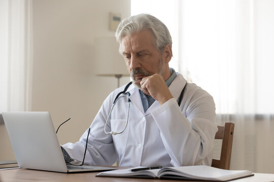 Pensive mature male doctor or GP sit at desk in hospital look at laptop screen thinking of patient diagnosis, thoughtful old man physician or therapist work on computer in cabinet, make decision
