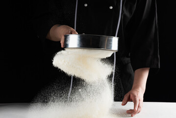 a dark background baker's hands sift white wheat flour over a sieve over