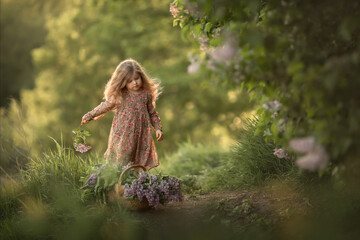 A little girl is standing near a large lilac Bush and playing with a basket filled with flowers. Image with selective focus and noise effect.
