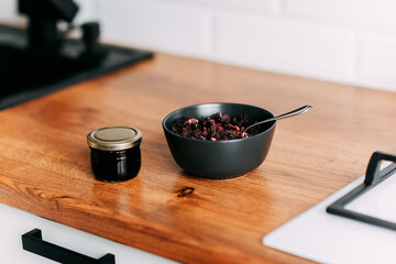 Loose tea in black bowl with spoon. Little honey can on the wooden table. Bright kitchen interior. Wooden complete kitchen with gas oven