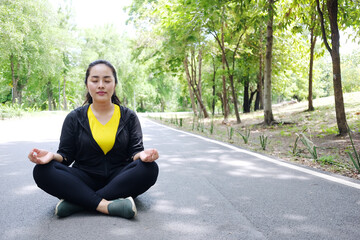 Smiling Asian young woman in sportwear close her eyes and sitting for doing yoga of meditation for create concentration and calm the mind on concrete floor after exercise in the natural light garden.